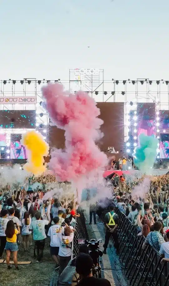 Crowd at an outdoor festival with colored smoke in front of the stage