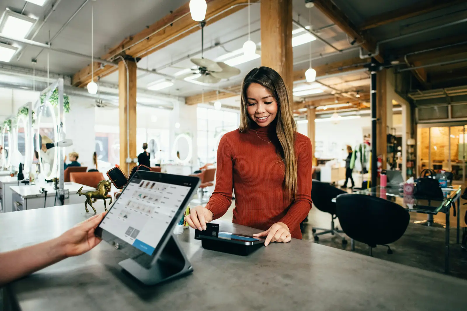 A smiling customer paying by card at a retail POS checkout counter while the clerk turns the tablet register toward her